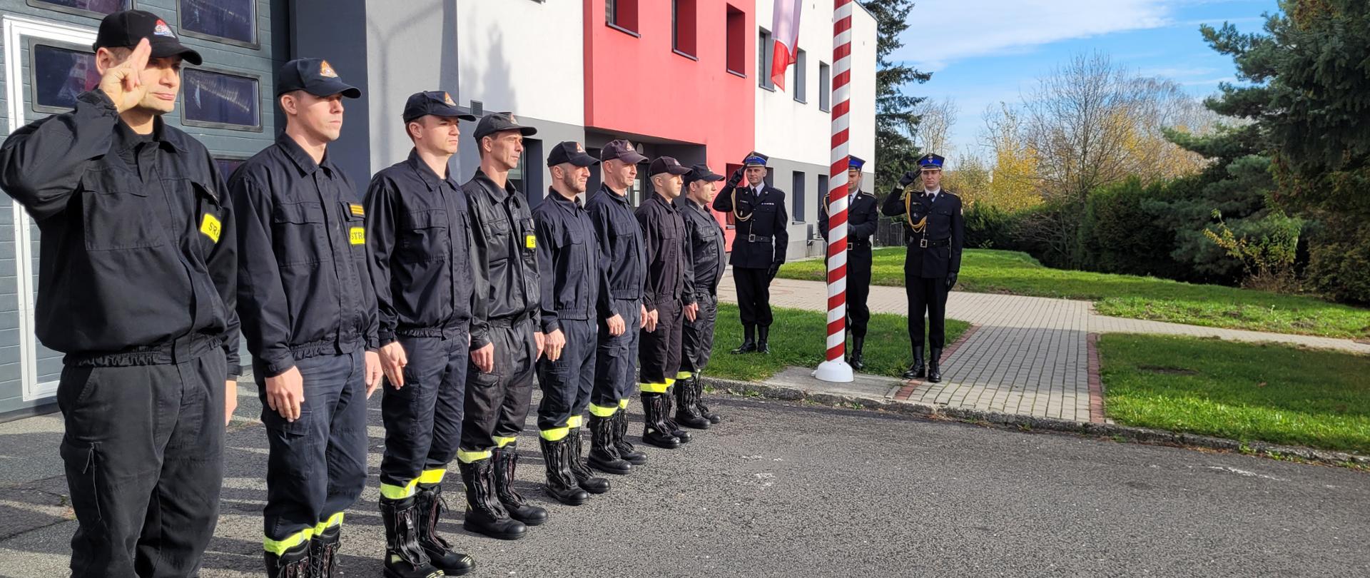 Strażacy stoją w szeregu przy maszcie flagowym. Poczet flagowy i dowódca salutują. Za strażakami budynek komendy w kolorach szaro - czerwonych. W prawej części zdjęcia skoszony trawnik i drzewo iglaste. 