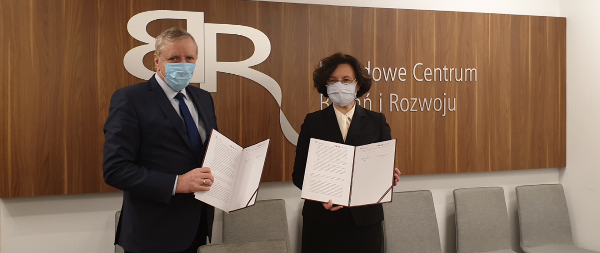 Na pierwszym planie, od lewej, stoją: dyrektor NCBR Wojciech Kamieniecki i dyrektor NAWA dr Grażyna Żebrowska. Trzymają w dłoniach otwarte teczki z treścią zawartego porozumienia. Elegancko ubrani, w maseczkach. W tle drewniana ścianka z logo Narodowego Centrum Badań i Rozwoju. Logo w kolorze srebrnym.