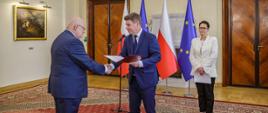 Minister Jan Grabiec wręcza akt powołania do Rady KSAP Janowi Pastwie, obok stoi szefowa służby cywilnej Anita Noskowska-Piątkowska; w tle flagi Polski i Unii Europejskiej