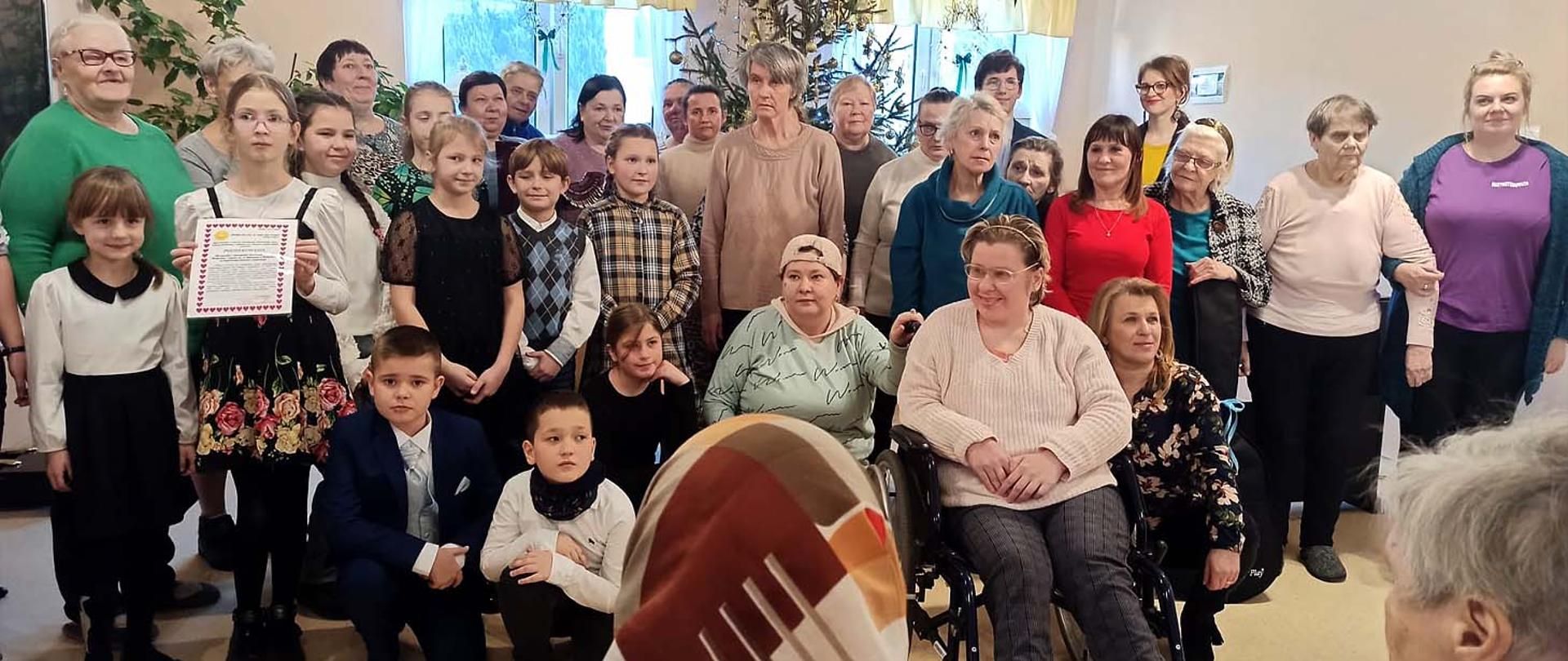 Na zdjęciu grupa kilkunastu osób, dorosłych i dzieci stoi w pokoju, część dorosłych z widoczną niepełnosprawnością. Za nimi ściana z oknem a przy niej udekorowana choinka.