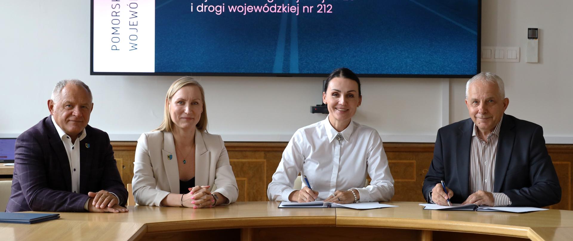 Cztery osoby siedzą przy stole konferencyjnym podczas podpisywania umowy. W tle wyświetlono informację o dofinansowaniu przebudowy drogi powiatowej nr 1770G w wysokości 1 276 449,38 zł.