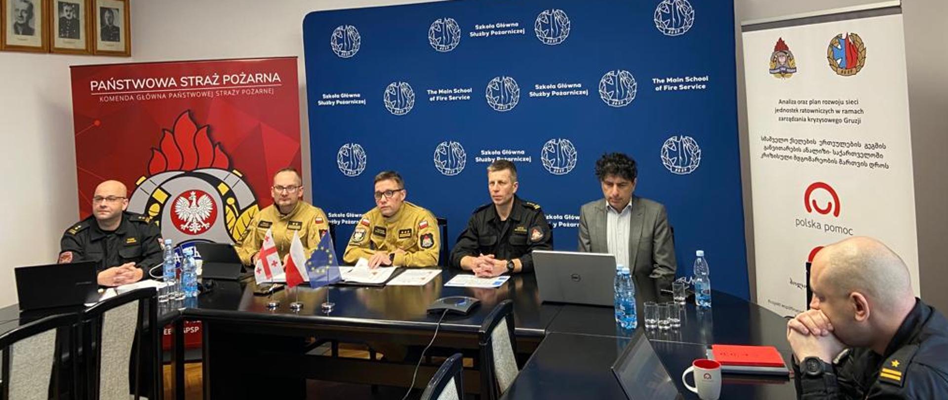 Pięciu mężczyzn w mundurach oraz jeden w garniturze siedzący przy stole w trakcie wideokonferencji. W tle ścianki z logotypami 