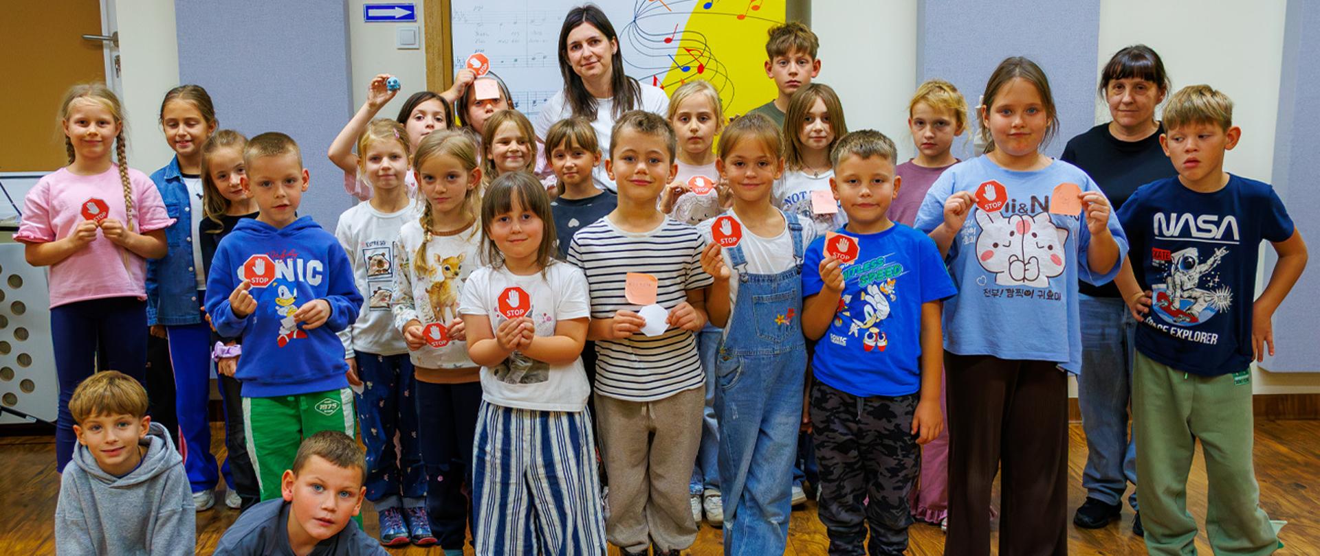 Grupa kilkanaścioro dzieci i nauczycielka stoją w auli szkolnej. W rękach trzymają znaczki z napisem STOP.