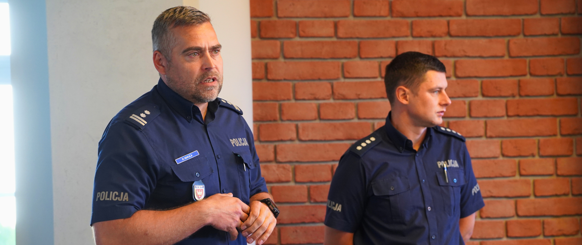 Policjanci prowadzą szkolenie dla strażaków, stojąc na sali przodem do zebranych, w tle prezentacja na telewizorze. 