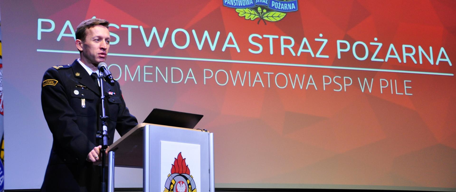 strażak stoi przy mównicy, za nim czerwone tło z logo państwowej straży pożarnej komenda powiatowa psp w pile