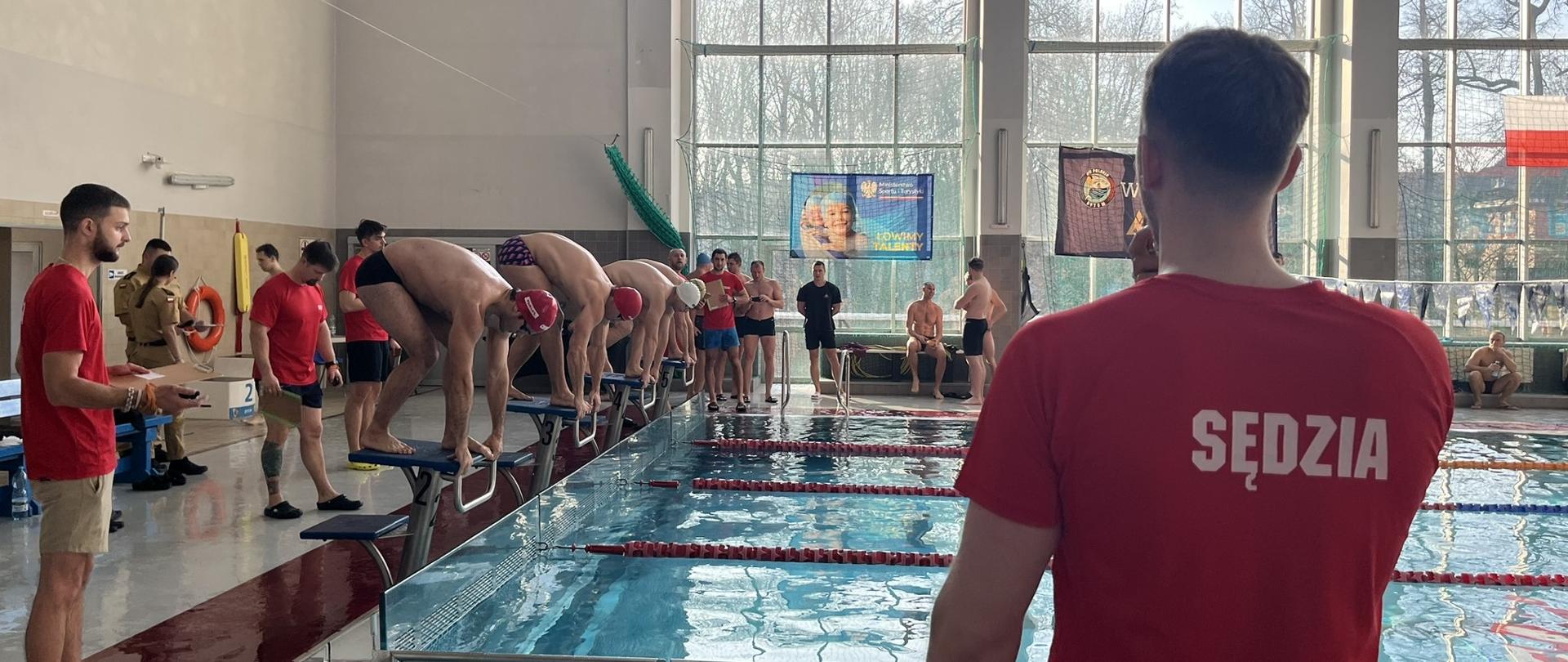 Na krytej pływalni kilku zawodników w strojach kąpielowych stoi pochylonych na blokach startowych, przygotowując się do rozpoczęcia wyścigu. Przy torach znajdują się osoby w czerwonych koszulkach, w tym jedna z napisem „Sędzia” na plecach, obserwująca start. W tle widać innych uczestników zawodów oraz duże okna, przez które wpada dzienne światło.
