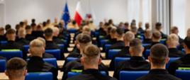 Zdjęcie od tyłu sali wykładowej – widoczni młodzi słuchacze PSP w czarnych mundurach siedzący w rzędach, skierowani w stronę stołu prezydialnego, przy którym siedzą oficerowie. W tle flagi Polski i Unii Europejskiej.