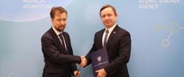 Na zdjęciu widać dwóch mężczyzn, którzy uścisnęli sobie dłonie. Mężczyźni ubrani są w ciemne garnitury, białe koszule i krawaty. Uśmiechają się i patrzą w kierunku aparatu. Mężczyzna po lewej stronie trzyma w dłoni granatową teczkę na dokumenty. Mężczyzna po prawej stronie również trzyma w dłoni ciemny folder, na którym widnieje godło Polski (biały orzeł).Za nimi znajduje się tło w jasnoniebieskim kolorze. Widać napisy: "Państwowa Agencja Atomistyki" oraz w języku angielskim: "National Atomic Energy Agency".