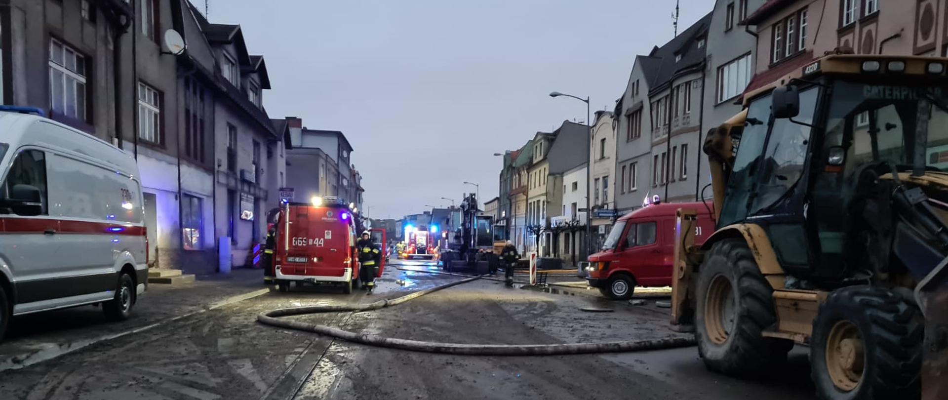Na zdjęciu widoczna ulica w mieście. Po obydwu stronach dwupiętrowe budynki mieszkalne. Na ulicy stoją koparki i samochody specjalne straży pożarnej i strażacy w ubraniach specjalnych. Na ulicy leżą węże szlamowe, odprowadzające wodę z zalanych przez deszcz budynków. 