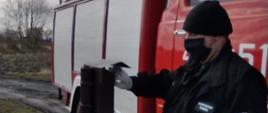 Zdjęcie przedstawia strażaka w czarnym mundurze koszarowym, który wkłada ulotki do skrzynki na listy. Strażak ma ubrane rękawiczki latexowe i czarna maseczkę. W tle czerwono-biało-srebrny samochód strażacki i zachmurzone niebo.

