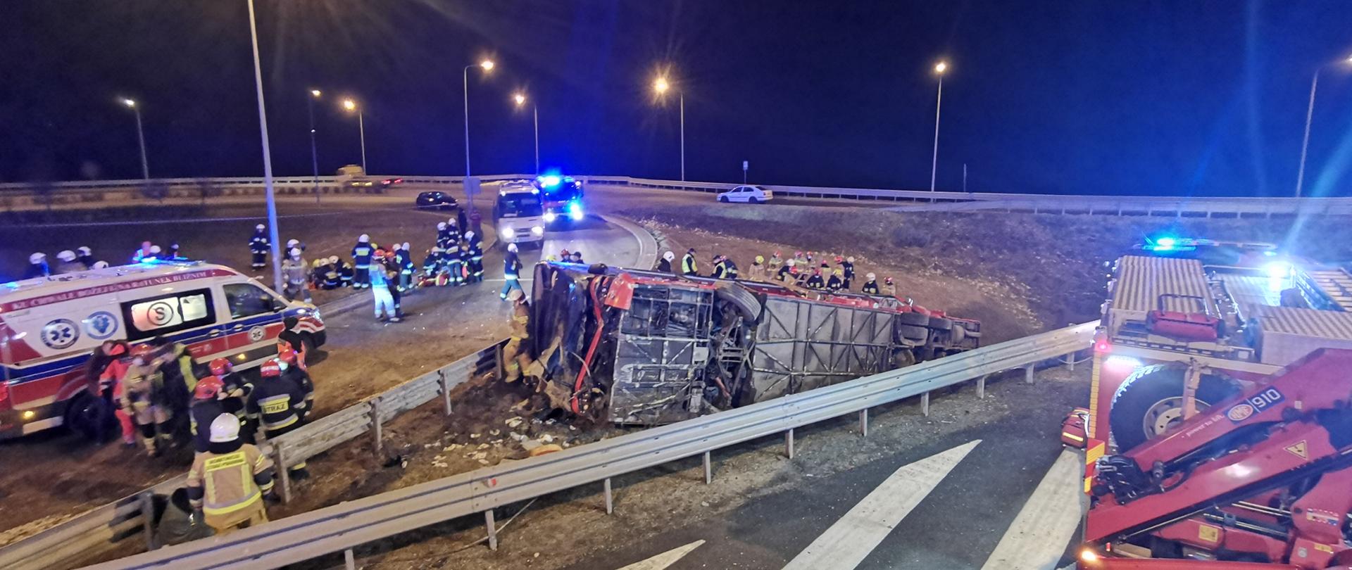 Na zdjęciu widać autobus przewrócony na prawy bok, leżący w rowie melioracyjnym przy autostradzie. Na jezdni widać pojazdy pożarnicze, ambulans oraz podstawiony bus. Na miejscu pracują strażacy, policjanci, a także ratownicy medyczni. Niedaleko od autobusu strażacy zajmują się kilkoma osobami poszkodowanymi.