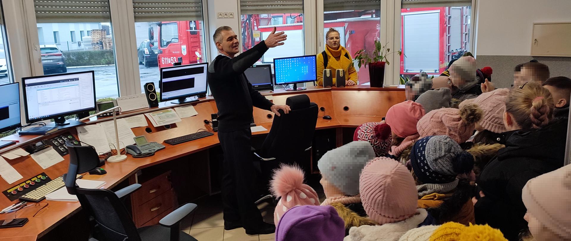 Grupa dzieci w pomieszczeniu Stanowiska Kierowania Komendanta Powiatowego PSP w Środzie Wielkopolskiej zapoznaje się ze specyfiką pracy Dyżurnego 