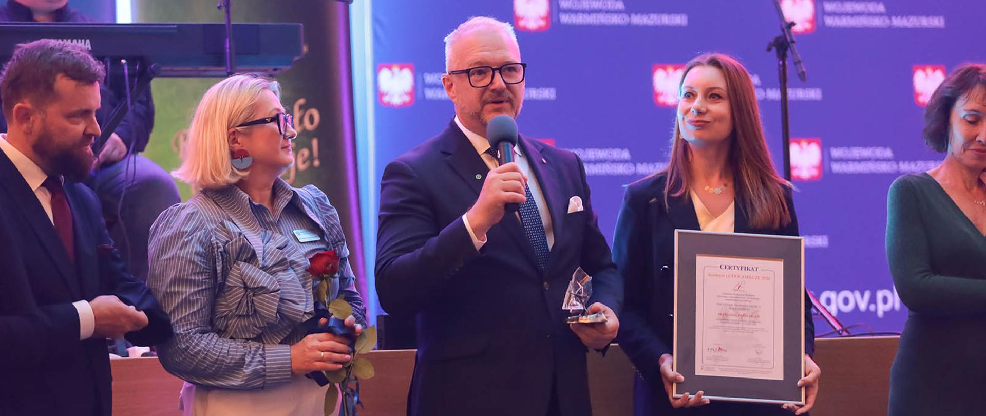 Mężczyzna w granatowym garniturze, wojewoda, przemawia przez mikrofon, kobieta w granatowej marynarce trzyma wyróżnienie wyróżnienie podczas wydarzenia 