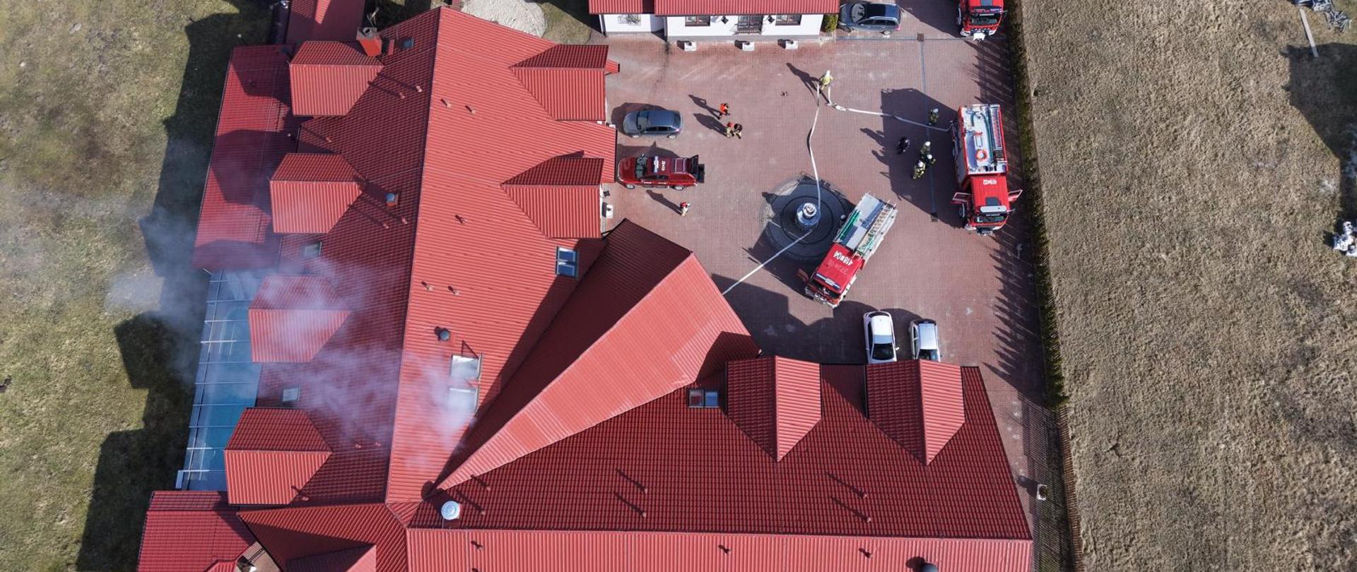 Rzut z pola widzenia drona z góry, na którym widać obiekt DPS, z którego wydobywa się zadymienie oraz strażaków i wozy bojowe.