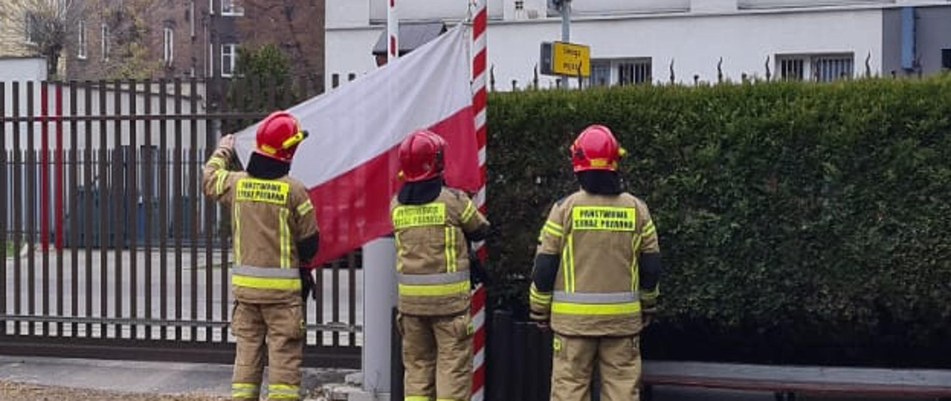 Zdjęcie przedstawia trzech funkcjonariuszy Państwowej Straży Pożarnej wieszających flagę Rzeczpospolitej Polskiej na maszt