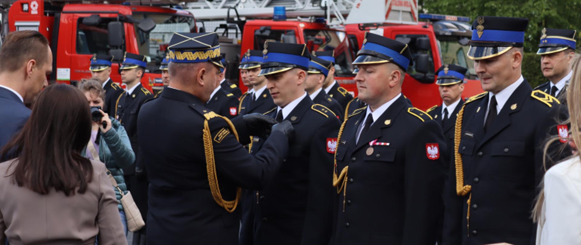 Zdjęcie zrobione na zewnątrz podczas uroczystości. Oficer Państwowej Straży Pożarnej w stopniu generała przypina funkcjonariuszowi Państwowej Straży Pożarnej medal. Za nimi stoją ustawieni strażacy Państwowej Straży Pożarnej wraz z pocztem sztandarowym. W tle stoją ustawione samochody ratowniczo-gaśnicze. 