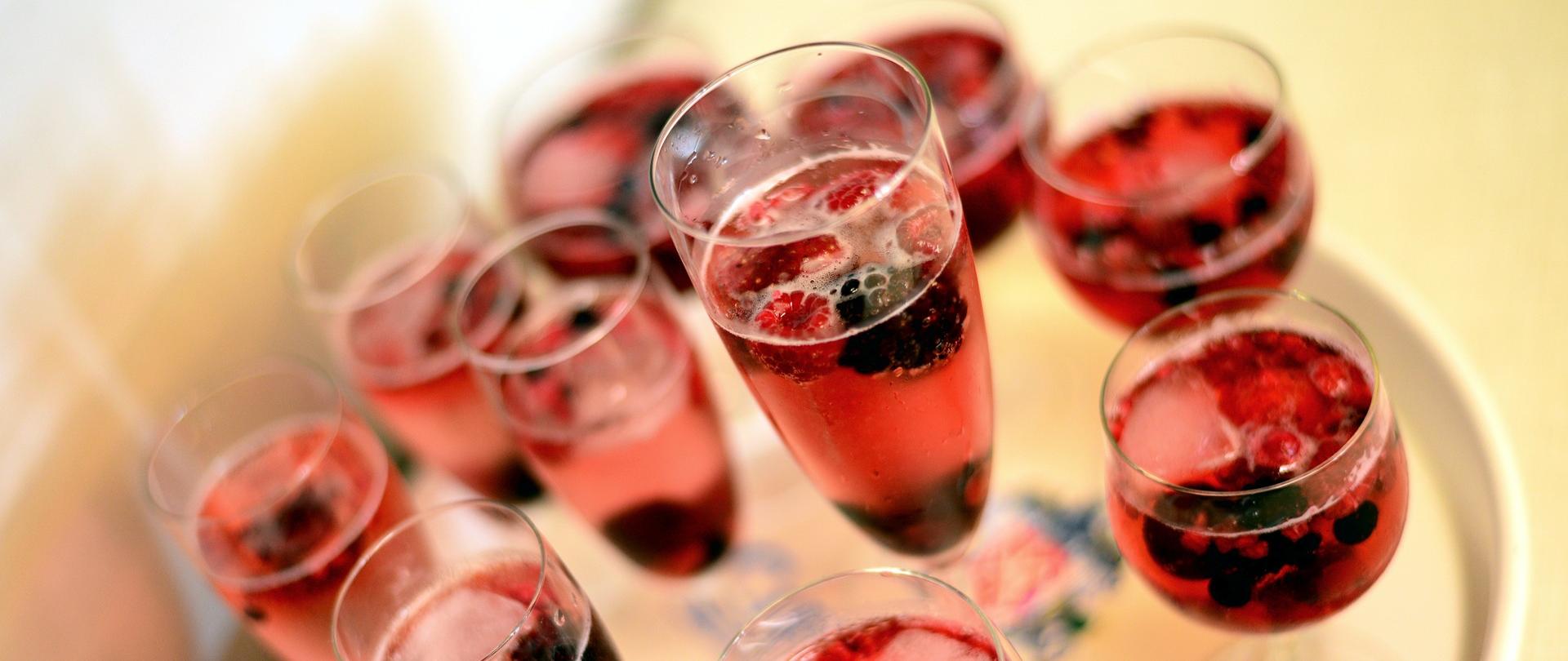 Na tacy stoją kieliszki z szampanem, w których znajdują się owoce: truskawki, maliny, jeżyny, czerwona porzeczka. 