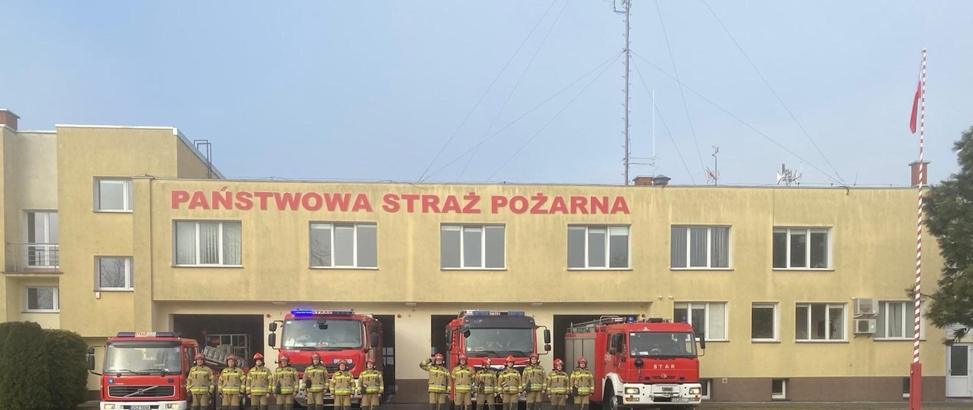 Pora dzienna. Funkcjonariusze KP PSP w Sztumie w pełnym umundurowaniu stoją przed bramami wyjazdowymi a za nimi wozy bojowe na sygnałach świetlnych.