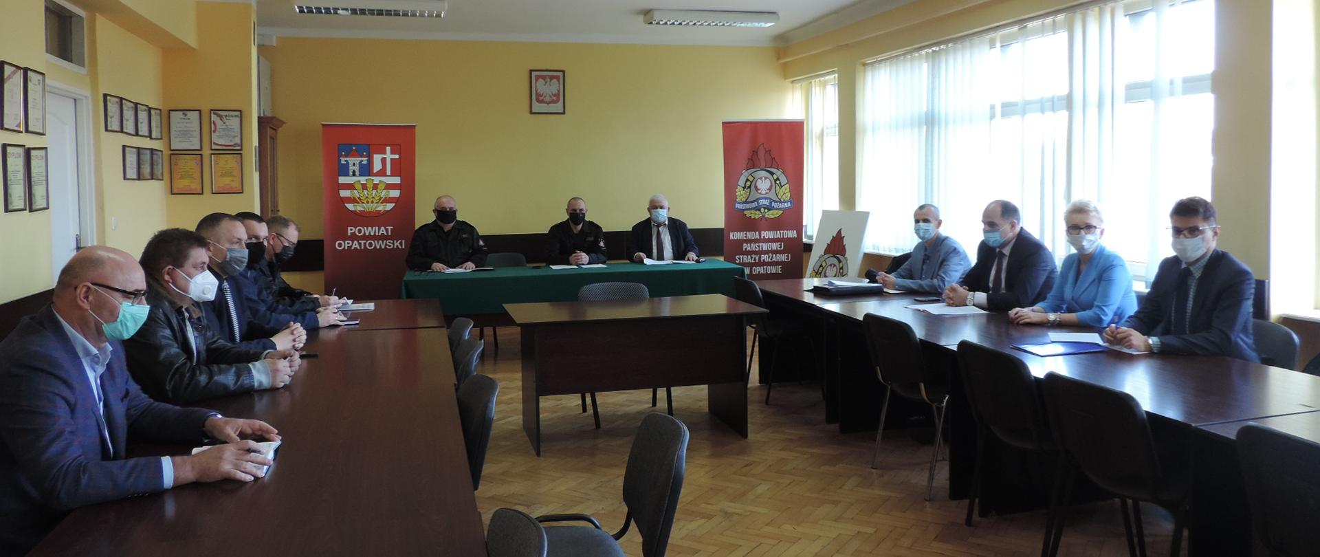 Zdjęcie przedstawia 12 osób siedzących przy stołach. Wśród tych osób znajduje się komendant powiatowy PSP w Opatowie oraz zastępca komendanta powiatowego oraz przedstawiciele jednostek samorządu terytorialnego. W tle dwa banery PSP
