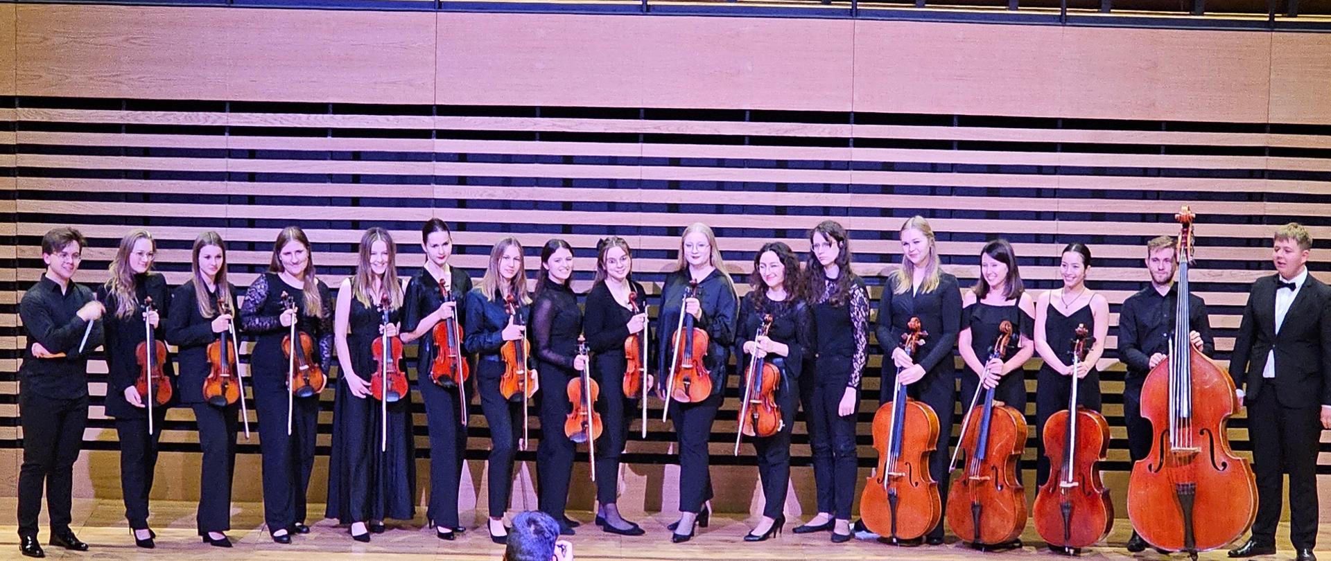 Siedemnaścioro osób stoi w rzędzie trzymając przed sobą skrzypce, altówki, trzy wiolonczele i jeden konrtabas.