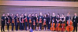 Siedemnaścioro osób stoi w rzędzie trzymając przed sobą skrzypce, altówki, trzy wiolonczele i jeden konrtabas.