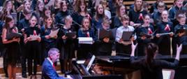 Około trzydzieścioro dzieci stojących w trzech rzędach śpiewa trzymając w rękach przed sobą nuty. Przed nimi mężczyzna gra na fortepianie, po prawej kobieta odwrócona tyłem do publiczności dyryguje.