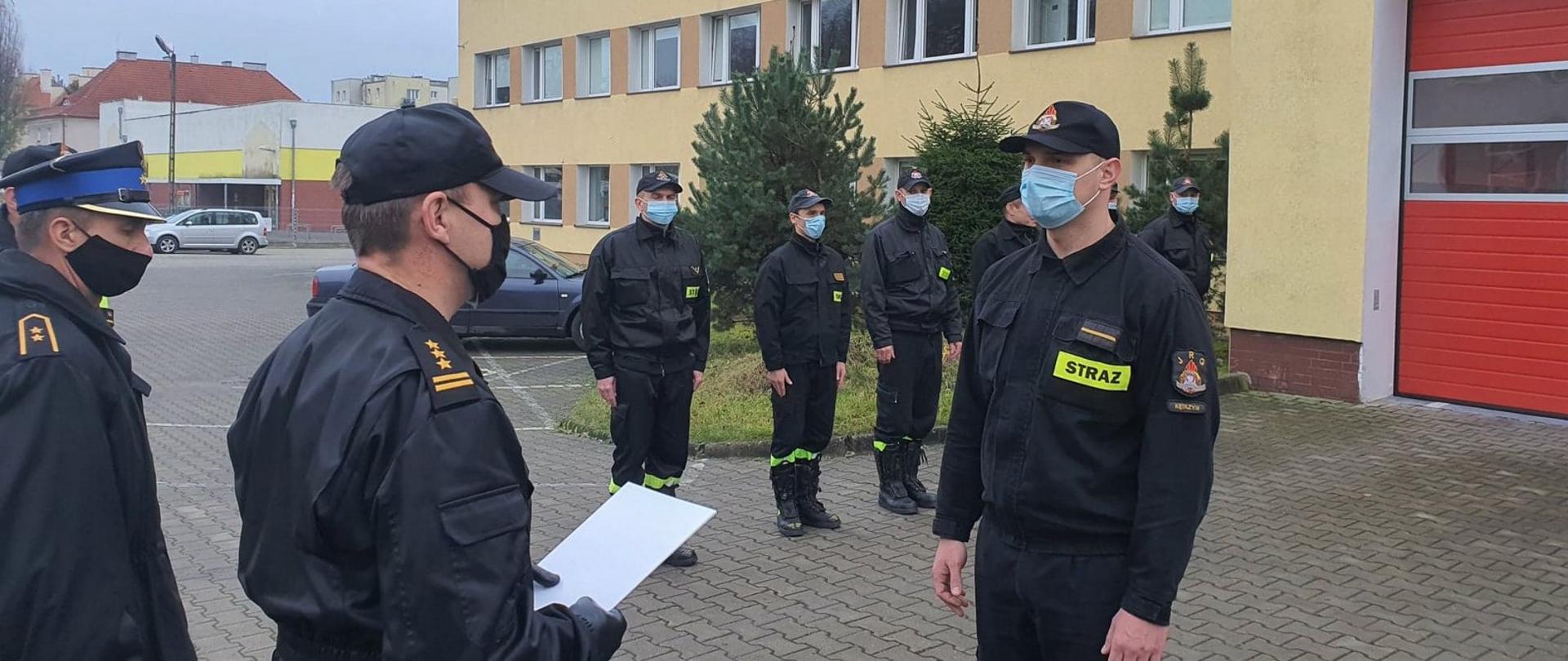 Zdjęcie zrobione na placu wewnętrzny KP PSP w Kętrzynie podczas uroczystej zbiórki podczas której odczytany został Wyciąg z Rozkazu Personalnego w sprawie nadania wyższych stopni. Strażacy ubrani w Koszar Dowódczo Sztabowy lub Ubranie Koszarowe. Komendant st. bryg. Krzysztof Grabias rozmawia z sekc. Dariuszem Trockim, w tle widać zmianę służbową ustawioną w szeregu.