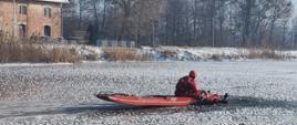 Dwóch strażaków znajduje się z dala od brzegu. Jeden ze strażaków znajduje się na plecach: częściowo na lodzie, częściowo w wodzie. Drugi strażak znajduje się w pozycji siedzącej na deskosaniach
i trzyma za ramiona drugiego strażaka wyciągając go z wody. Strażacy ubrani w kombinezony wypornościowe koloru czerwonego, zabezpieczeni linkami ratowniczymi.
Tafla lodu pokryta płatkami śniegu, w tle widoczne drzewa, stary budynek i barierki mostu.