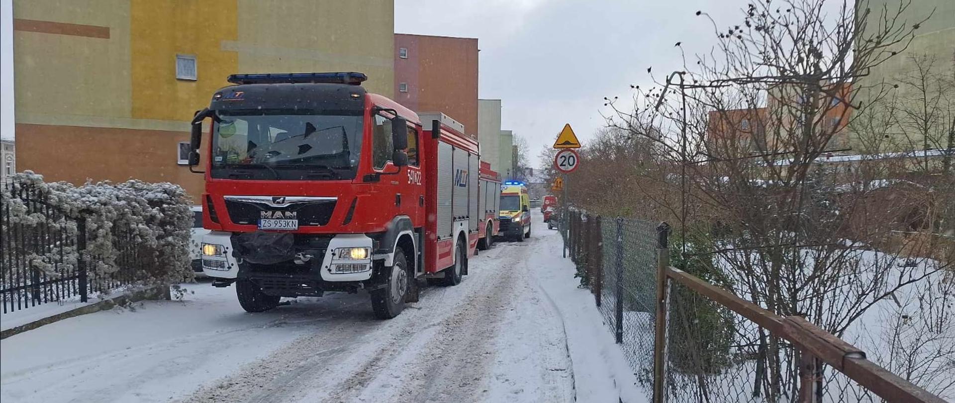 Czerwony wóz strażacki i ambulans stoją na wąskiej, ośnieżonej ulicy między blokami mieszkalnymi; w tle widoczne są budynki, ogrodzenia i znak ograniczenia prędkości do 20 km/h.