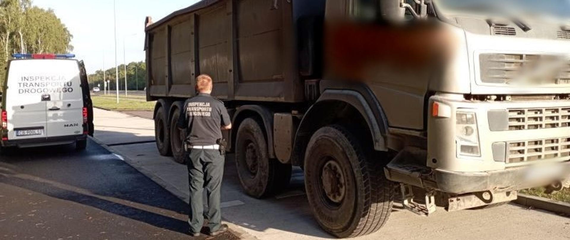 Na pierwszym planie: przód i prawa strona wywrotki zatrzymanej do kontroli drogowej przez patrol kujawsko-pomorskiej ITD. Obok ciężarówki stoi umundurowany inspektor bydgoskiej ITD. W tle (od lewej strony): tył oznakowanego furgonu ITD.