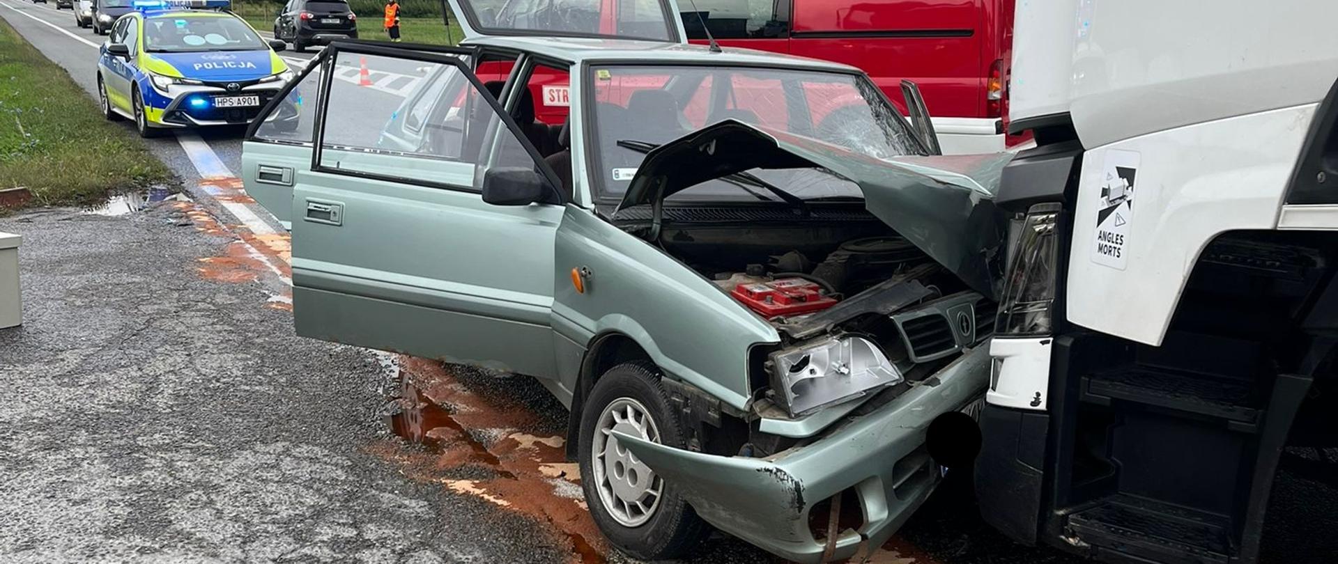 Widok z przodu na uszkodzony, samochód osobowy marki Polonez, który uderzył w przód białego ciągnika siodłowego. Auto osobówki ma zniszczoną maskę oraz prawą stronę przodu, otwarte drzwi kierowcy i pasażera oraz podniesioną klapę bagażnika. Na jezdni widoczny jest wyciek płynów eksploatacyjnych w kolorze rdzawym. W tle stoją pojazdy służb: radiowóz policyjny oraz czerwony wóz strażacki z oznaczeniem 409 40.