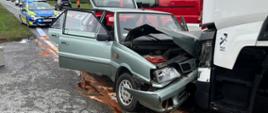 Widok z przodu na uszkodzony, samochód osobowy marki Polonez, który uderzył w przód białego ciągnika siodłowego. Auto osobówki ma zniszczoną maskę oraz prawą stronę przodu, otwarte drzwi kierowcy i pasażera oraz podniesioną klapę bagażnika. Na jezdni widoczny jest wyciek płynów eksploatacyjnych w kolorze rdzawym. W tle stoją pojazdy służb: radiowóz policyjny oraz czerwony wóz strażacki z oznaczeniem 409 40.