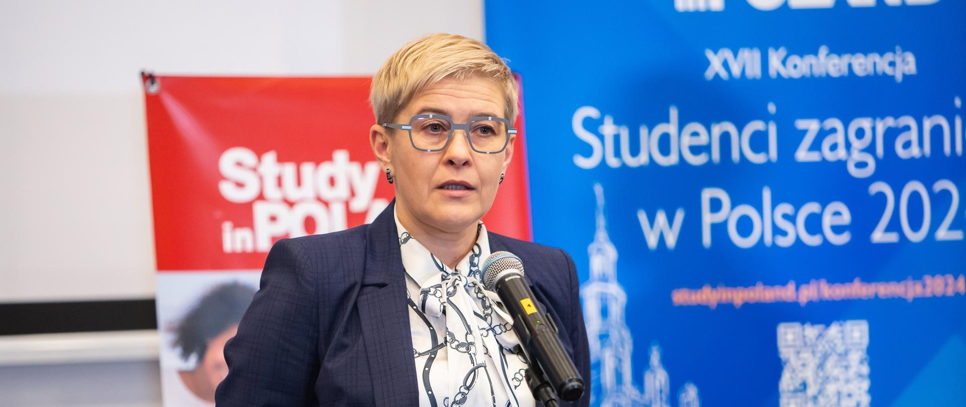 Wiceminister Mrówczyńska stoi i mówi do mikrofonu, za nią na ścianie ekran z napisem Studenci zagraniczni w Polsce 2024.
