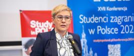 Wiceminister Mrówczyńska stoi i mówi do mikrofonu, za nią na ścianie ekran z napisem Studenci zagraniczni w Polsce 2024.