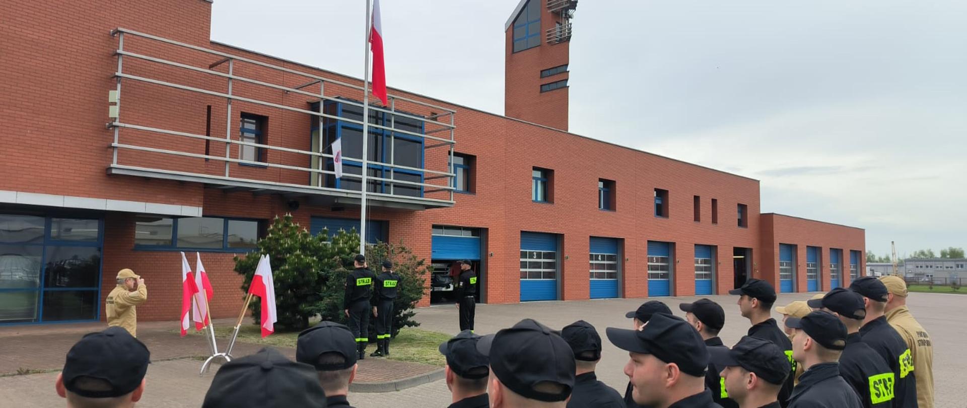 Poczet flagowy podczas wciągania flagi na maszt, obok komendant i w tle strażacy stojący w dwuszeregu