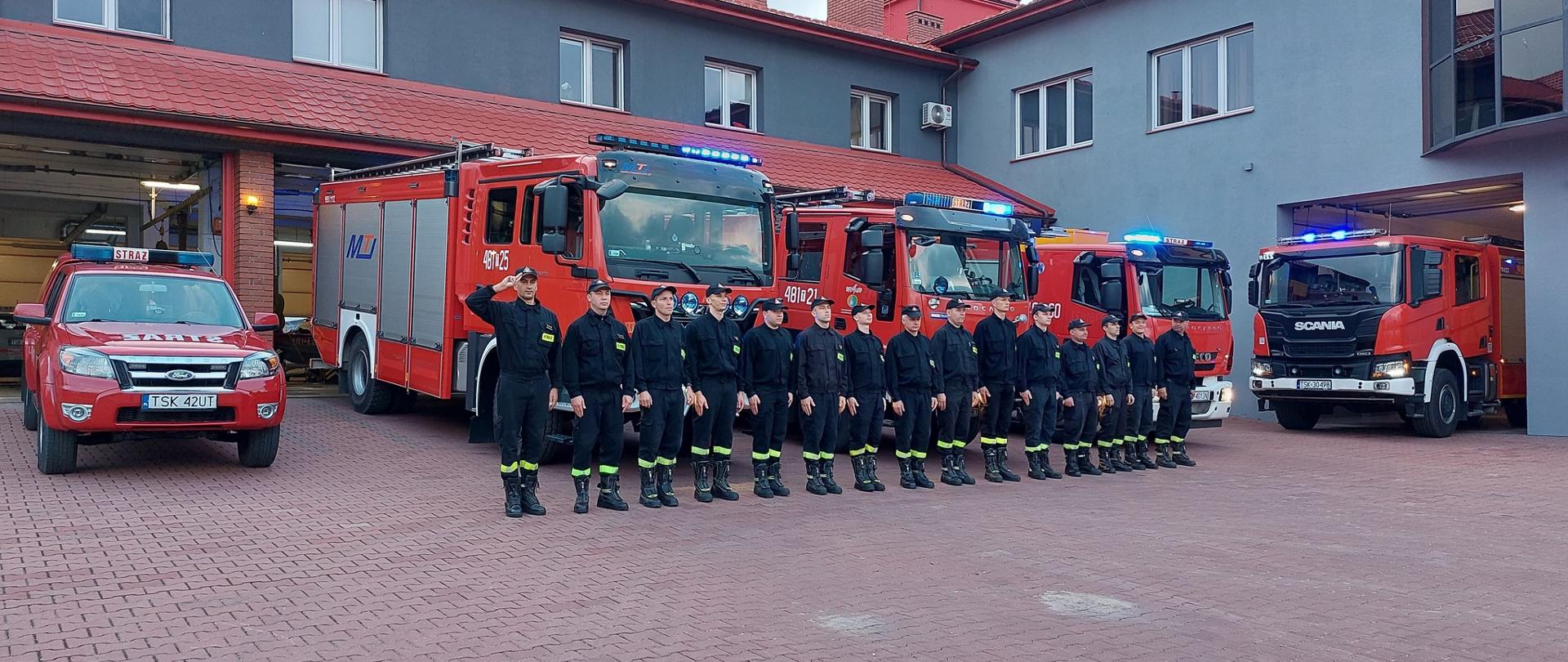 Strażacy na zbiórce przed komendą na tle samochodów z włączonymi sygnałami świetlnymi.
