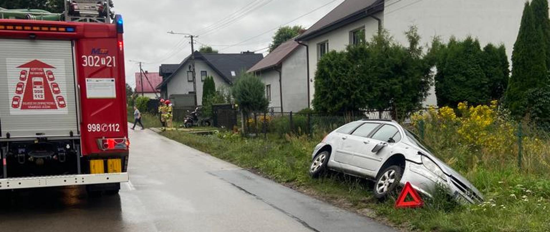 Zdjęcie przedstawia drogę powiatową gdzie doszło do wypadku drogowego. Po lewej stronie, na pasie jezdni stoi samochód pożarniczy, po drugiej stronie drogi, w przydrożnym rowie, na boku leży samochód osobowy. 