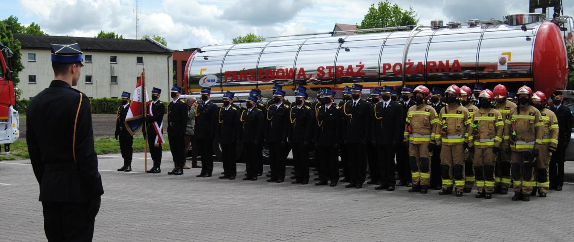 Na pierwszym planie znajduje się grupa strażaków ustawiona w szeregu, naprzeciw ich znajduje się prawdpodobnie dowóca uroczystości. Po lewej stronie szeregu większość strażaków ubrana jest w mundury galowe, dodatkowo widoczny jest poczet sztandardowy. Po prawej stronie widać strażaków ubranych w ubrania specjalne.
Pozują na tle cysterny z napisem "Państwowa Straż Pożarna".
