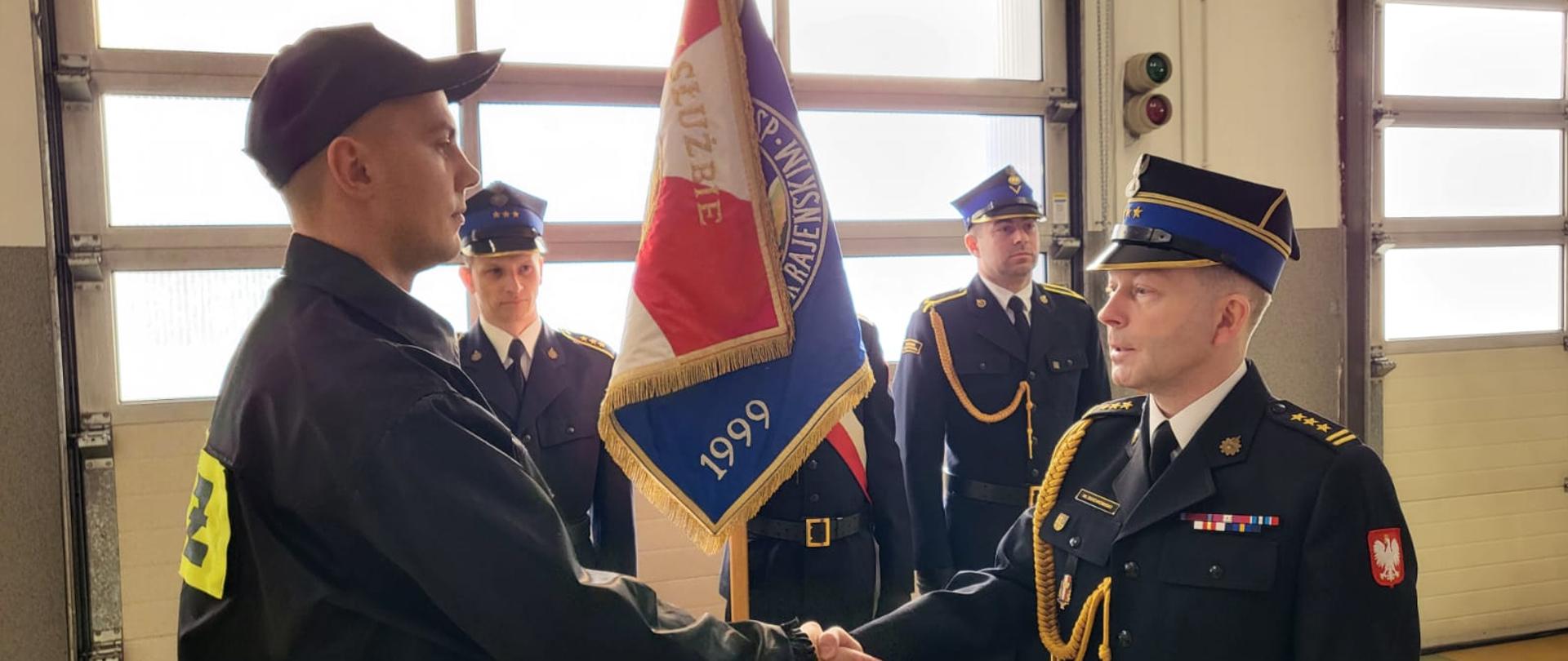 Starszy brygadier Maciej Suchomski ubrany w mundur wyjściowy gratuluje młodemu strażakowi ubranemu w mundur koszarowy, ściskając mu dłoń. W tle poczet sztandarowy Komendy Powiatowej Państwowej Straży Pożarnej w Sępólnie Krajeńskim.