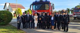 Zaproszeni goście wraz z przedstawicielami jednostki OSP Zabawa stoją w rzędzie przed samochodem strażackim, samochód przyozdobiony jest w biało czerwoną szarfę