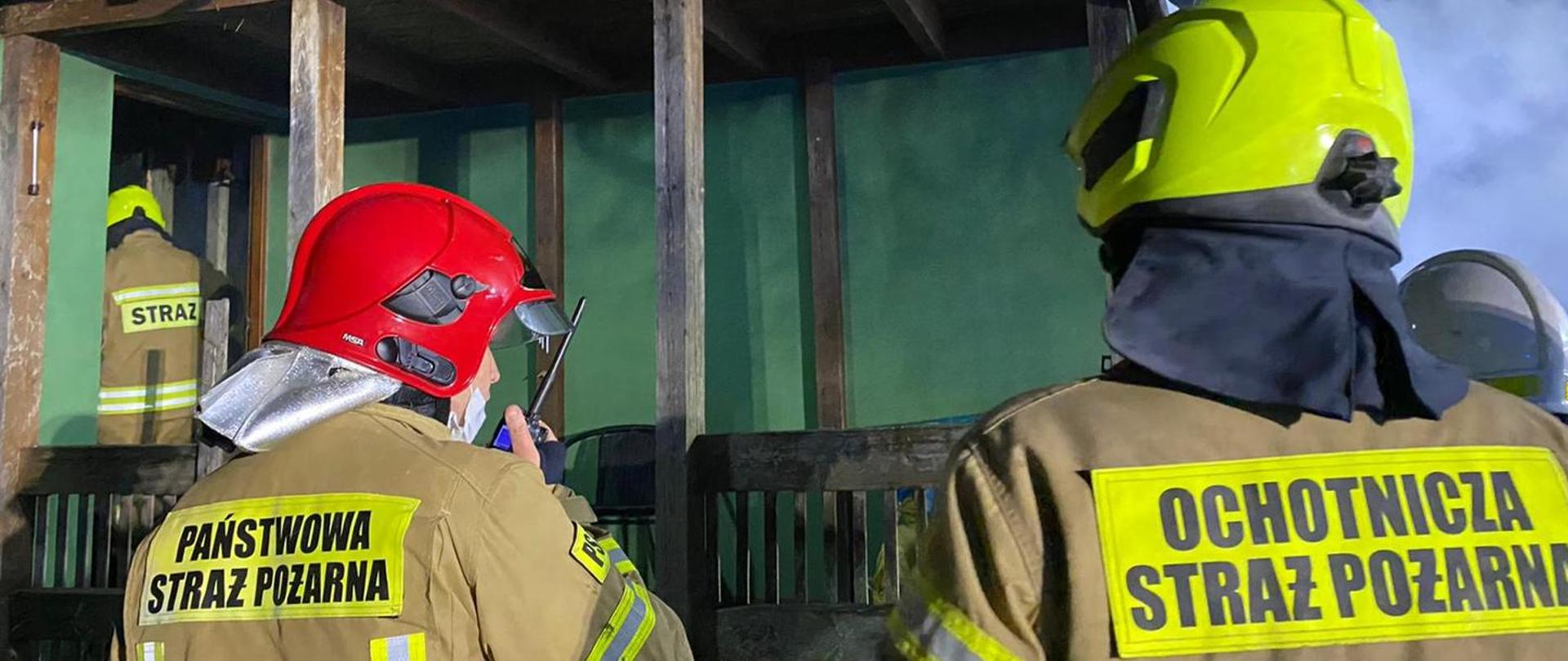 Zdjęcie przedstawia dwóch strażaków w umundurowaniu specjalnym stojących tyłem jeden ze strażaków prowadzi korespondencję radiową przez radiotelefon nasobny. Strażak z lewej strony ma na plecach napis Państwowa Straż Pożarna, a strażak z prawej strony Ochotnicza Straż Pożarna