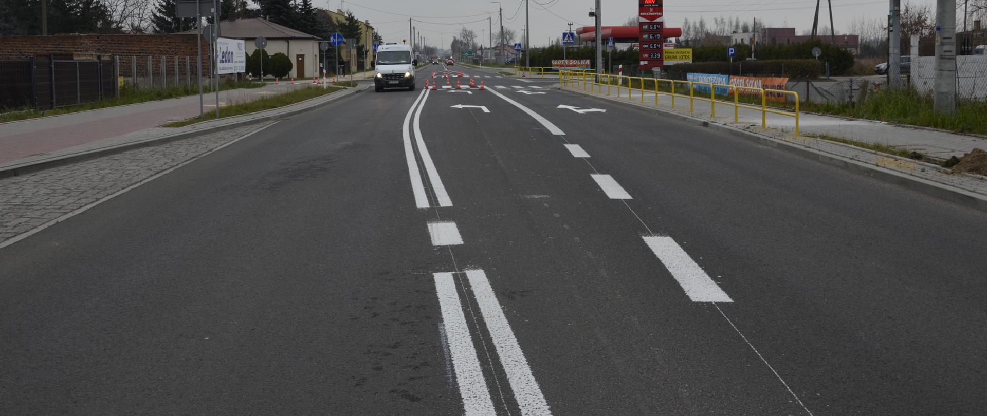 Widok na wyremontowaną drogę w kierunku południowym. Po lewej stronie częściowo widoczna zatoczka autobusowa, na wprost skrzyżowanie z ulicą podporządkowaną i wydzielonym pasem do skrętu w lewo, po prawej dwie stacje paliw. 