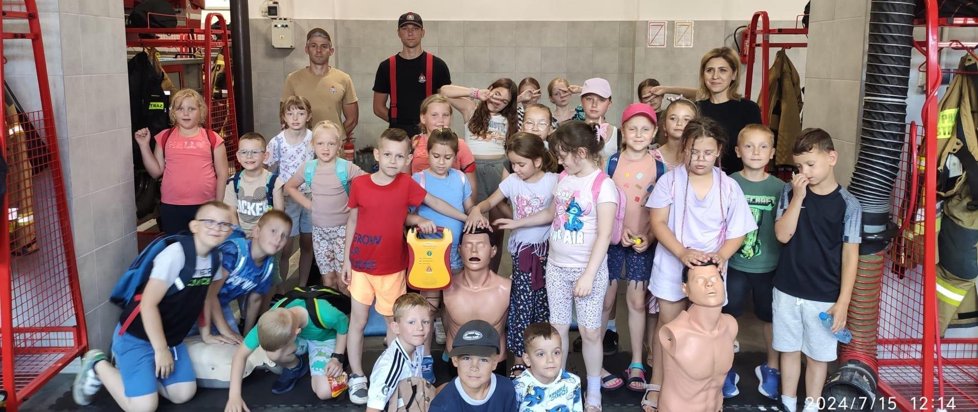 Grupa dzieci ze strażakami pozuje do zdjęcia w garażu bojowym.