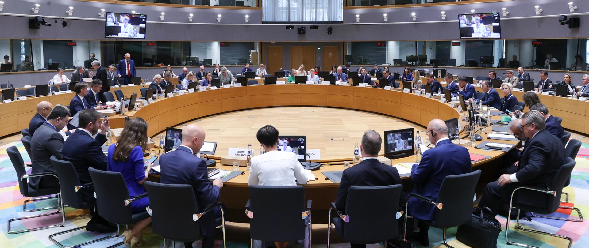 In the Europa building at the Council of the EU, in a modern conference room, the meeting participants are seated around a large, round table. In front of the delegations are monitors, microphones, documents and nameplates indicating the countries. In the background, further workstations and interpreters’ booths can be seen. The distinctive, multicoloured ceiling gives the interior a modern look.