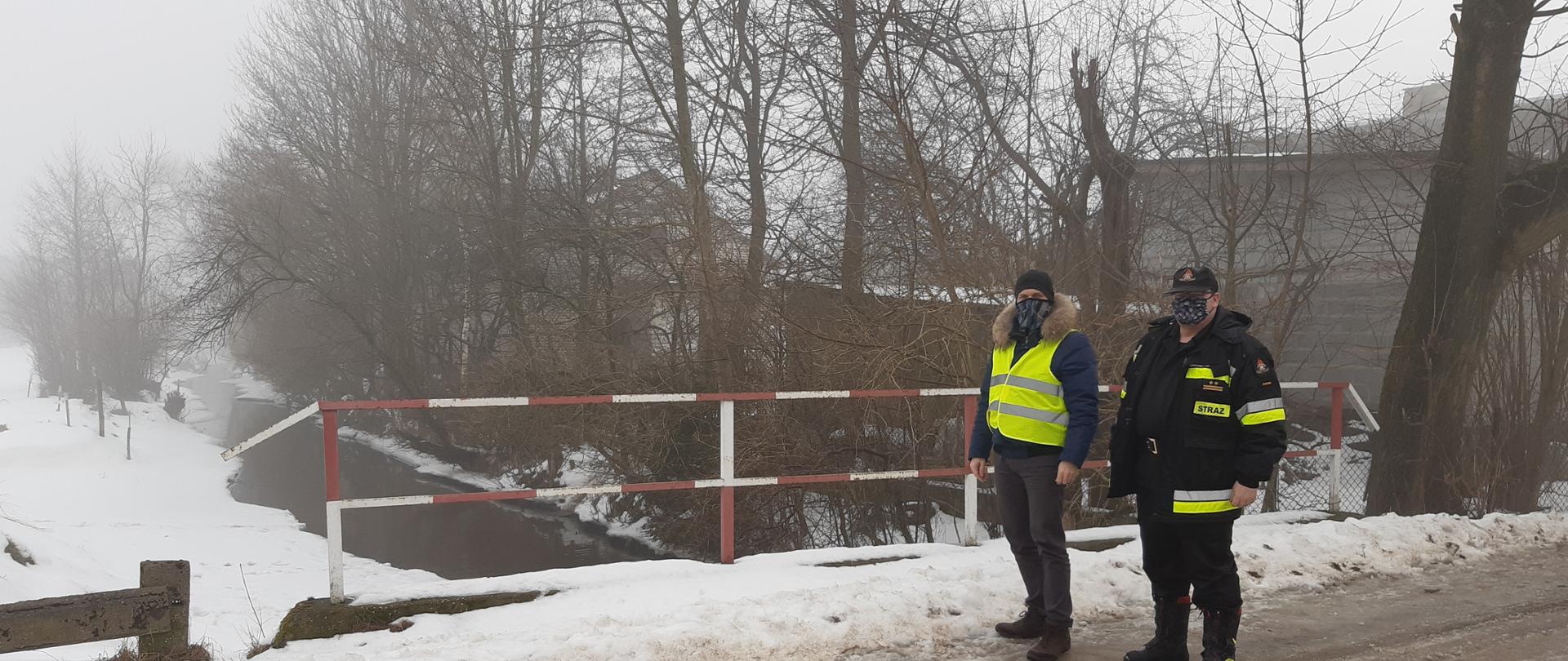Dwóch mężczyzn w maseczkach stojących przy barierkach zabezpieczających. W tle widać drzewa i strumyk wodny.