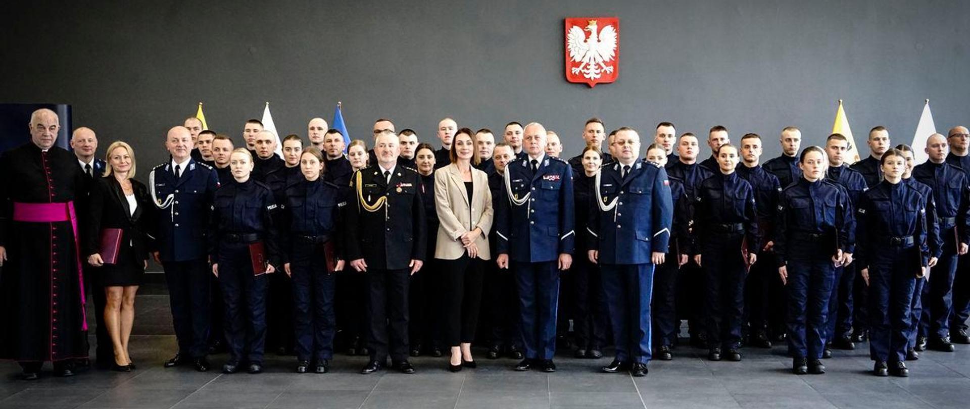 Kilkanaście osób stoi w dużej sali, na ścianie wisi godło