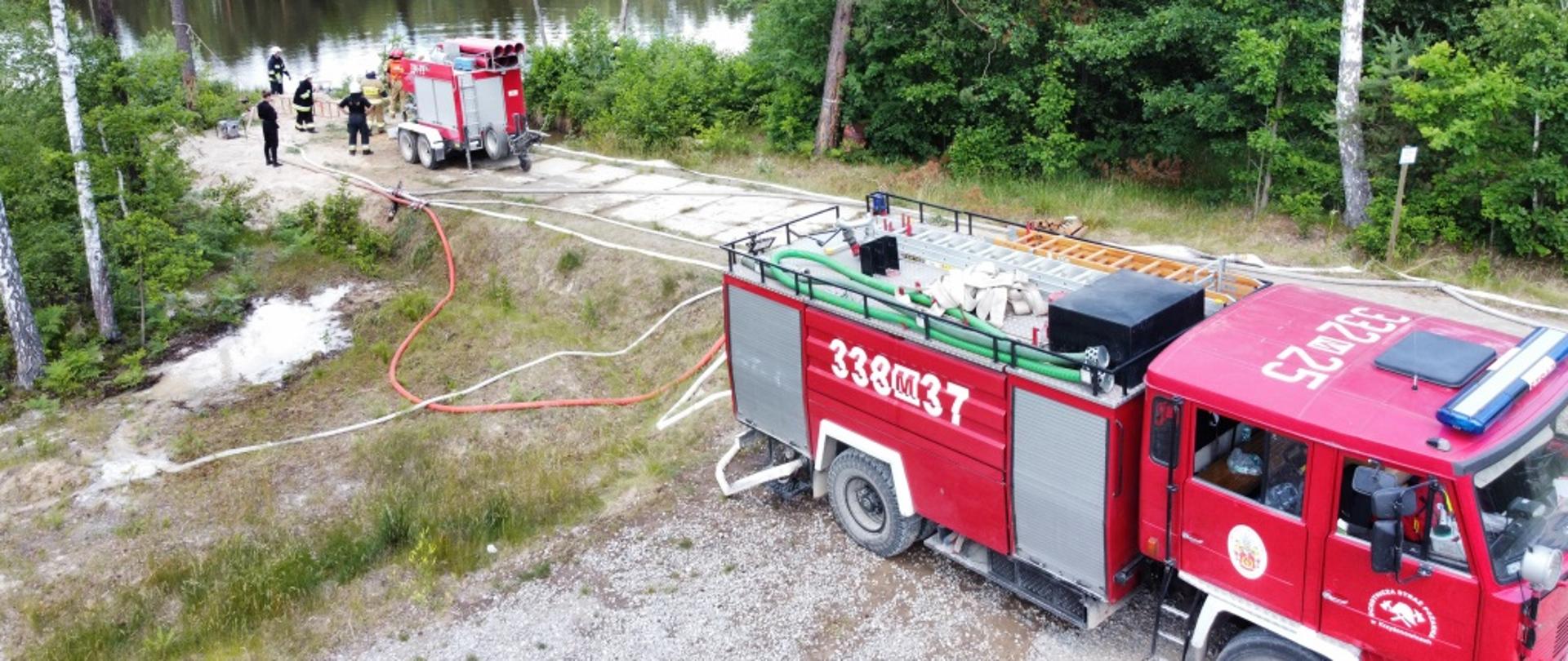 Zdjęcie wykonane przy pomocy drona na terenie Leśnictwa Jasieniec. Na zdjęciu widać zbiornik wodny nad brzegiem, którego ustawiona jest pompa dużej wydajności. Od pompy w prawą stronę, drogą szutrową ułożona jest magistrala z węży pożarniczych. Dookoła pompy widać również strażaków ubranych w odzież specjalną którzy obsługują punkt czerpania wody.