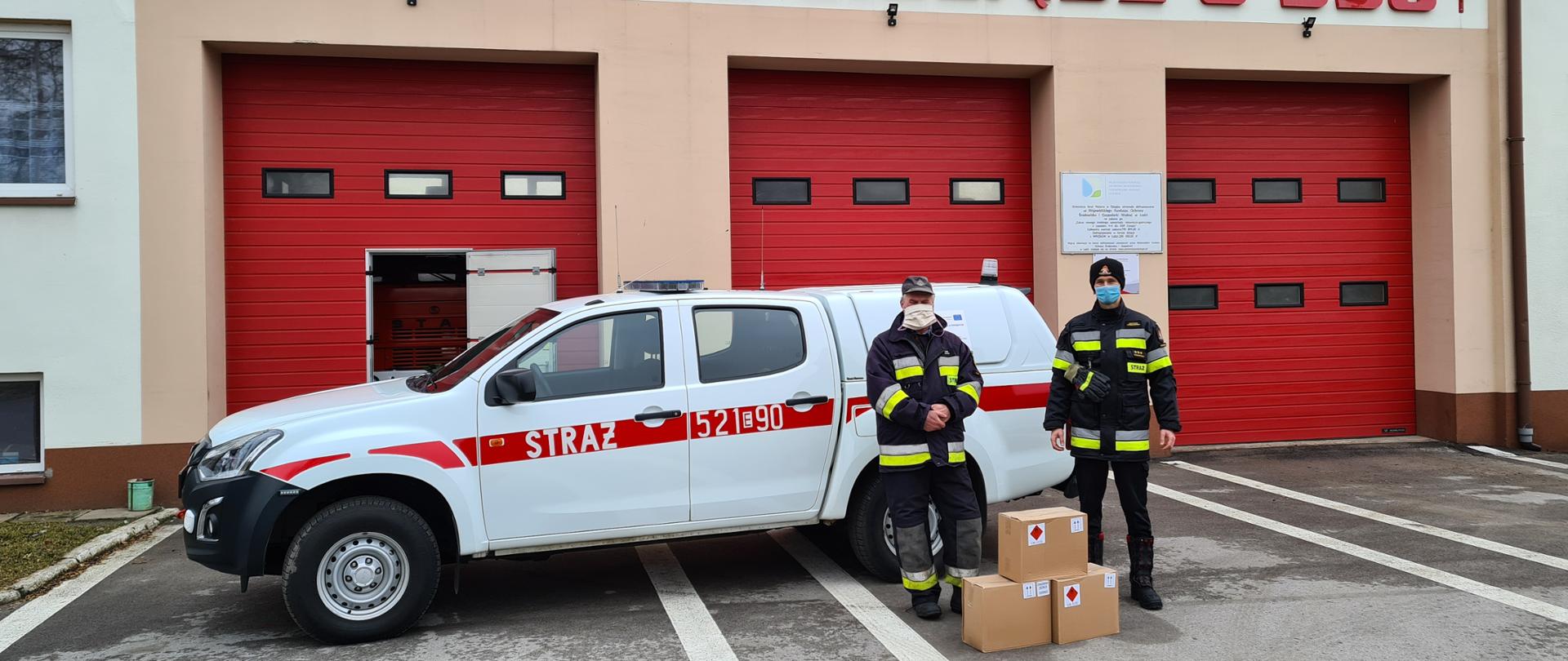 Na zdjęciu widać dwóch mężczyzn stojących na tle samochodu strażackiego typu SUV w kolorze białym, za którym znajduję się część budynku remizy OSP Cielądz. Budynek jest beżowy z czerwoną bramą garażową. Jeden z mężczyzn to ochotnik z OSP, ubrany jest w stój strażacki w kolorze granatowym, a drugi to strażak PSP Rawa Mazowiecka, ubrany jest w stój strażacki - granatowy/czarny. Między nimi stoją 3 pudła z środkami DO DEZYNFEKCJI.