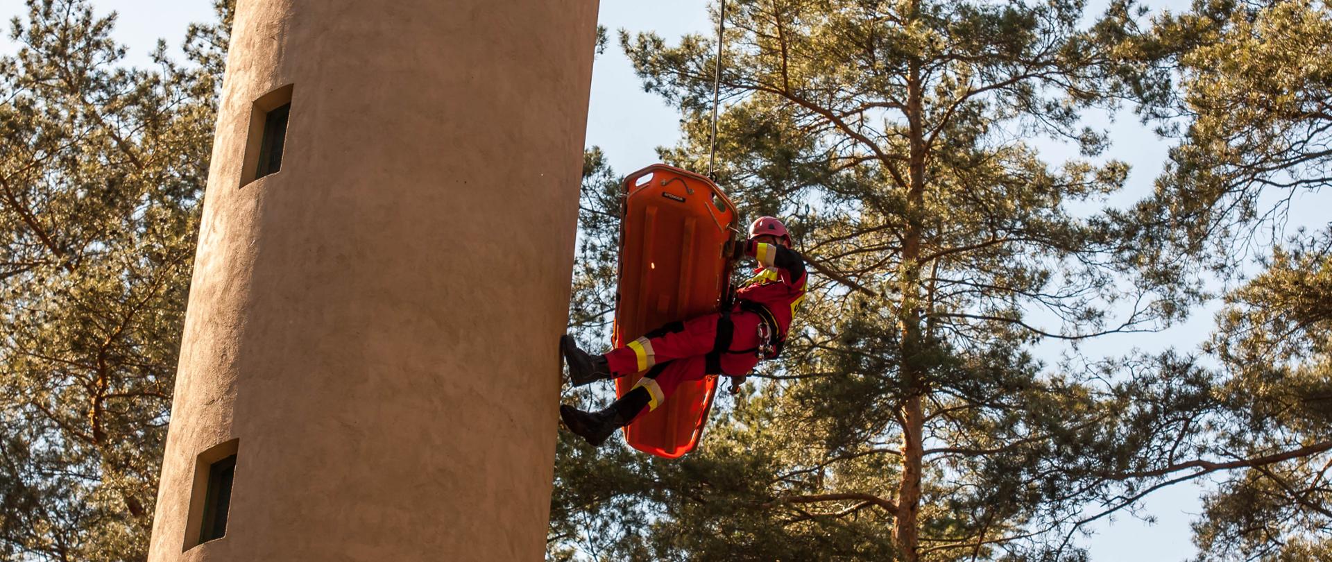 Na zdjęciu widoczny strażak Specjalistycznej Grupy Ratownictwa Wysokościowego "Głuchołazy" będący na wysokości połowy wieży dostrzegalni pożarowej (przy pomocy technik linowych) z podwieszonym na koszu ratowniczym koloru pomarańczowego poszkodowanym (manekin). Strażak ubrany w kombinezon koloru czerwonego, kask koloru czerwonego oraz rękawiczki. W tle widoczne korony drzew/