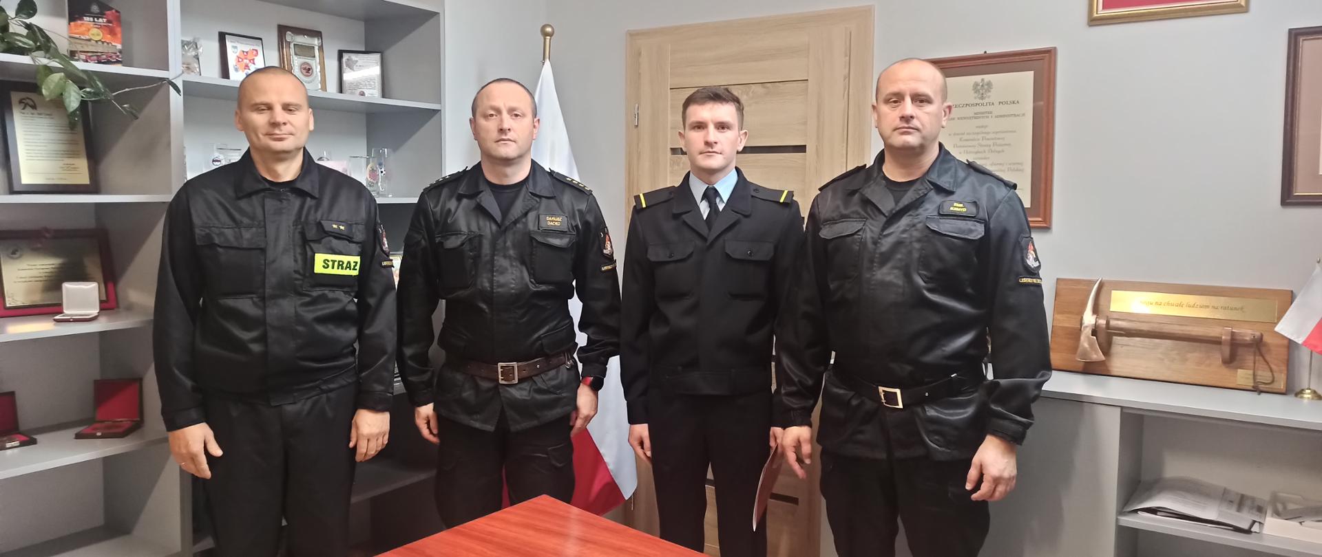 Na zdjęciu widać strażaka któremu wręczono akt mianowania do służby stałej, dowódcę zmiany, dowódcę jednostki ratowniczo gaśniczej oraz komendanta powiatowego PSP w Ustrzykach Dolnych. Zdjęcie wykonane jest w biurze komendanta. 
